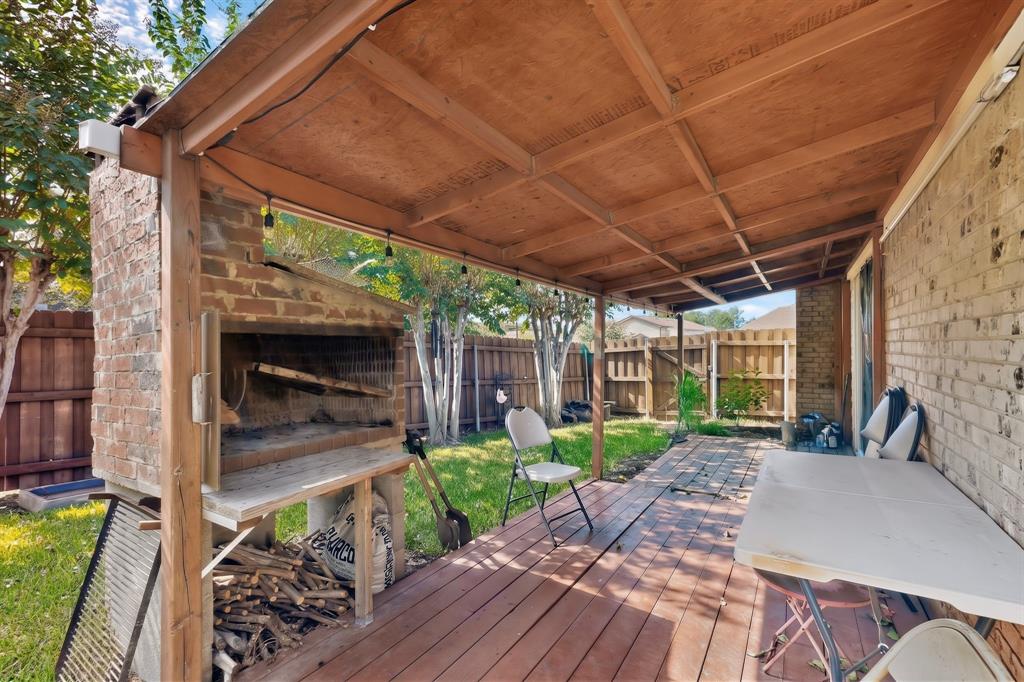 Covered wooden deck featuring a built-in masonry outdoor fireplace, exposed wood-beam ceiling, and brick exterior wall