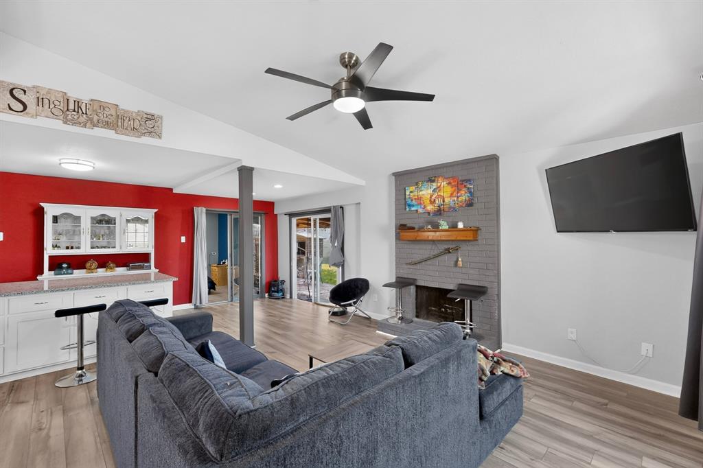 Living area featuring wood-finish flooring, vaulted ceilings, a grey painted brick fireplace with a wood mantel, a ceiling fan with integrated lighting, and a built-in wet bar with cabinetry Living area featuring wood-finish flooring, vaulted ceilings, a grey painted brick fireplace with a wood mantel, a ceiling fan with integrated lighting, and a built-in wet bar with cabinetry