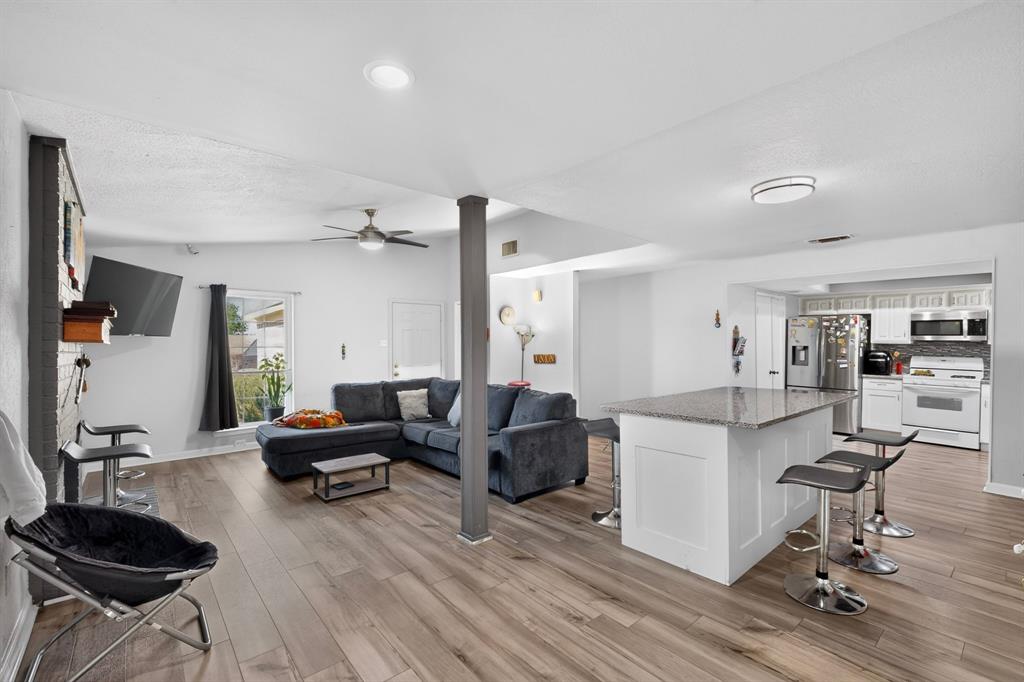 Open-concept interior featuring wood-finish flooring, a kitchen island with stone-finish countertop, white cabinetry, a structural support column, and a brick-faced fireplace surround
