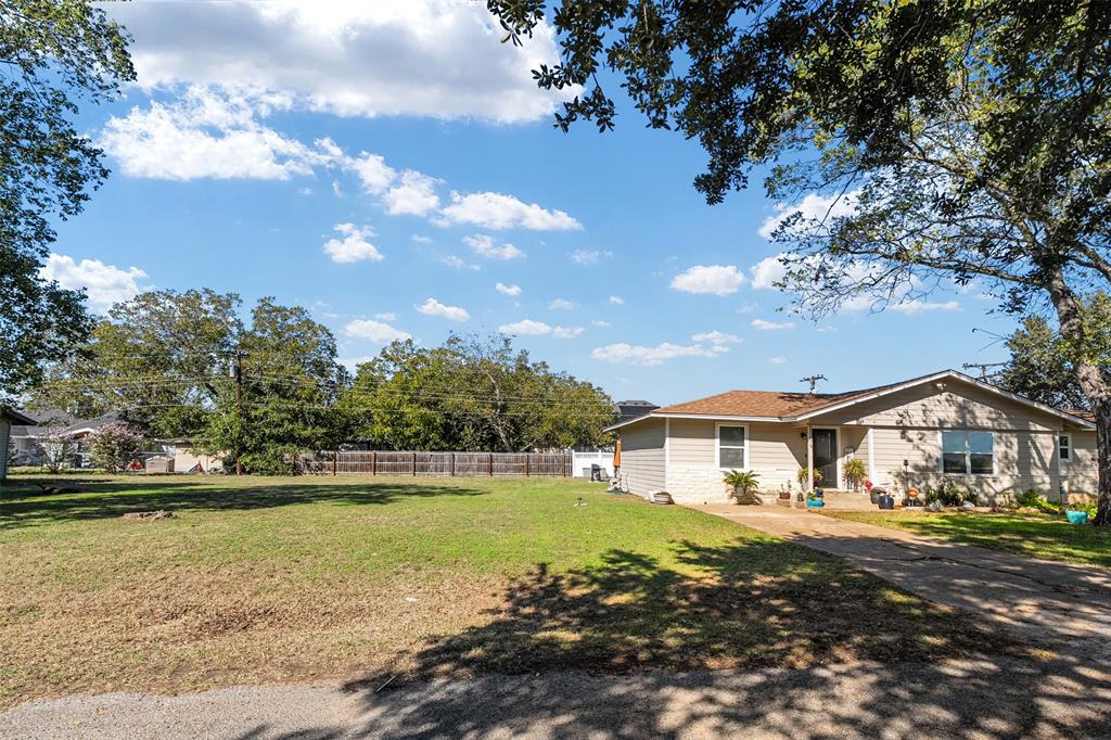 Exterior of a single-story residence featuring light-toned horizontal siding, a low-pitched roof, concrete driveway, and expansive front lawn with mature trees