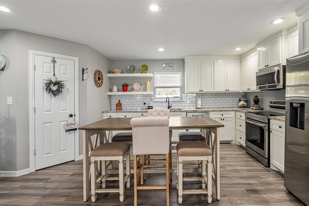 Kitchen featuring white shaker-style cabinetry, subway tile backsplash, stainless steel appliances, open floating shelving, and wood-finish flooring Kitchen featuring white shaker-style cabinetry, subway tile backsplash, stainless steel appliances, open floating shelving, and wood-finish flooring