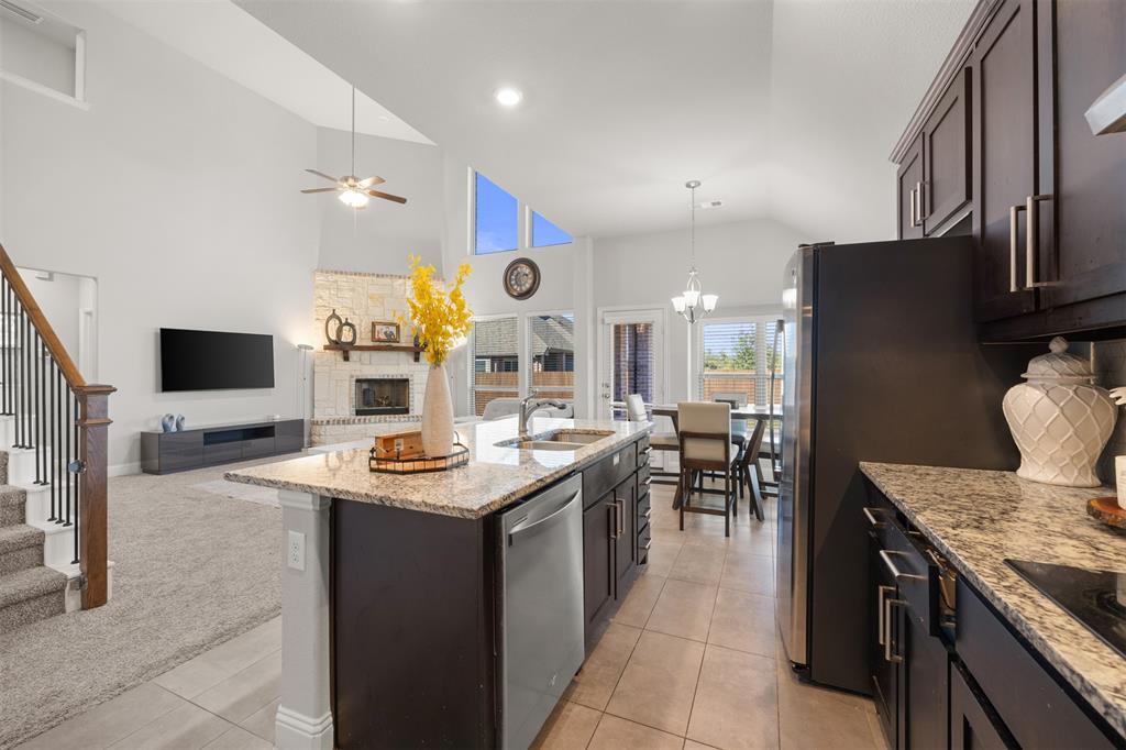 Open-concept kitchen featuring dark-finish cabinetry, stone-patterned countertops, stainless steel appliances, a center island with a built-in sink, and vaulted ceilings
