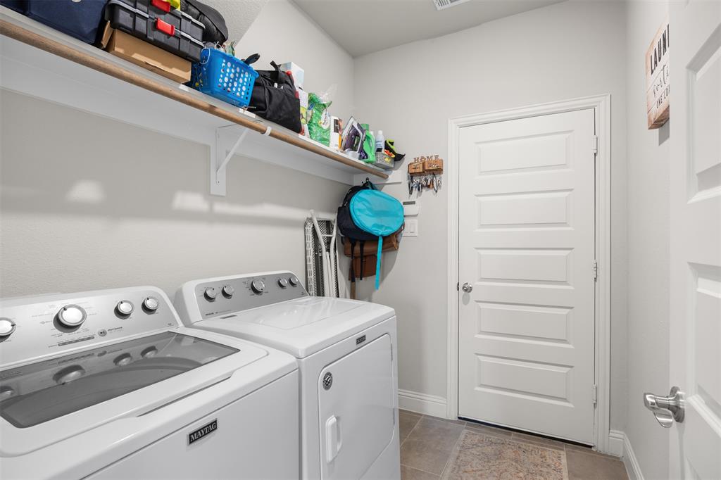 Laundry area featuring neutral-tone ceramic tile flooring, a multi-panel interior door with brushed metal hardware, and full-width wall-mounted shelving