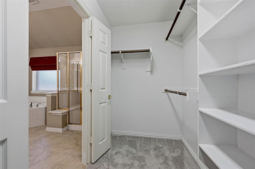 Walk-in closet featuring white built-in shelving and dual hanging rods, with tiled flooring and a glass-enclosed shower visible beyond the doorway