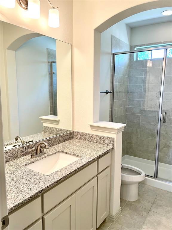 Bathroom featuring a stone-composite countertop with an integrated basin, shaker-style cabinetry, framed wall mirror, arched architectural detailing, and a walk-in shower with tile surround