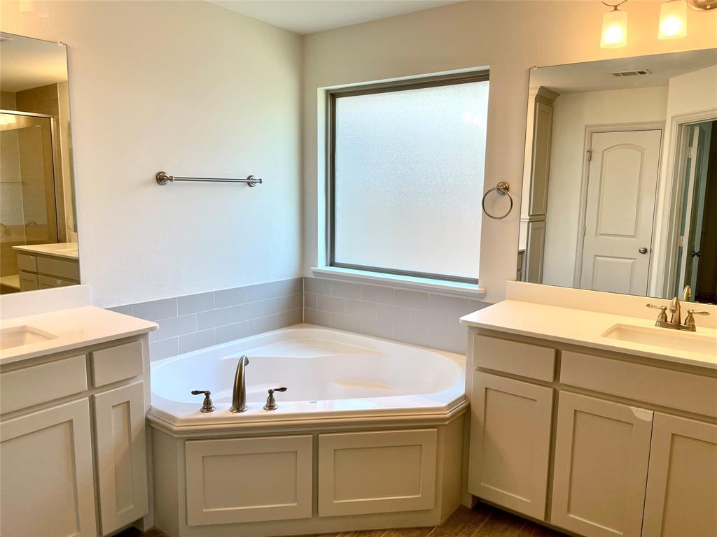 Corner soaking tub with tile surround, dual vanities featuring light-tone cabinetry, surface-mounted mirrors, and chrome fixtures