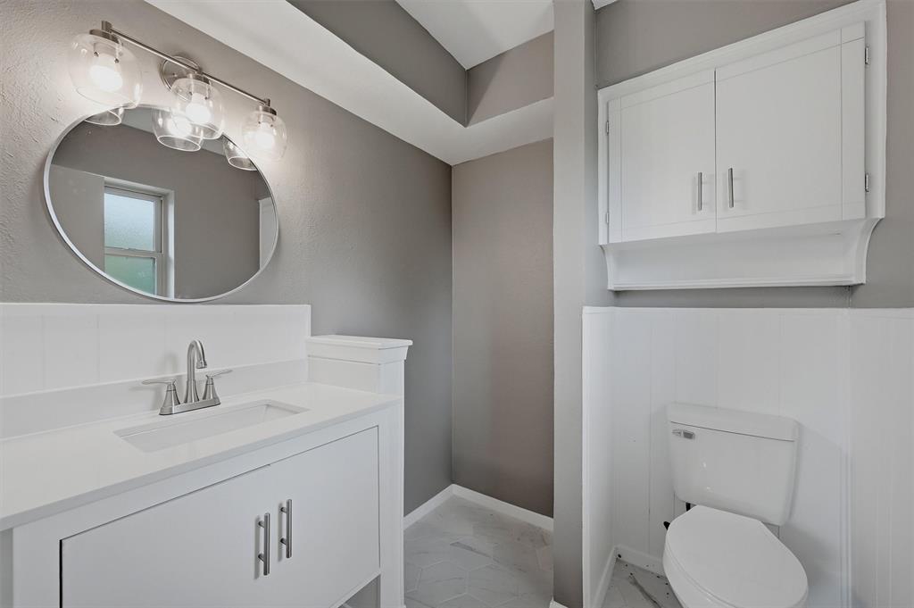 White vanity with rectangular undermount sink, oval wall-mounted mirror, three-bulb vanity light fixture, white wall-mounted storage cabinetry, and half-wall wainscoting