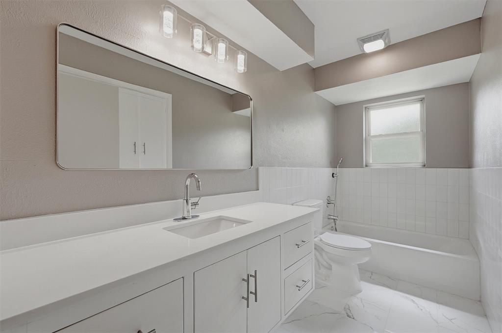Bathroom featuring a white vanity with integrated rectangular sink and stone-finish countertop, large wall-mounted rectangular mirror, four-light vanity fixture, white tile tub surround, and tile flooring