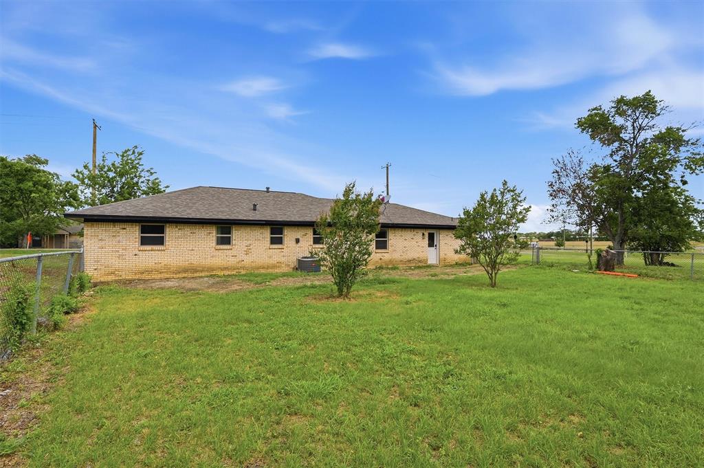 Exterior view featuring a tan brick facade, hip roof with composition shingles, metal chain-link perimeter fencing, and mature trees on a level grass lot
