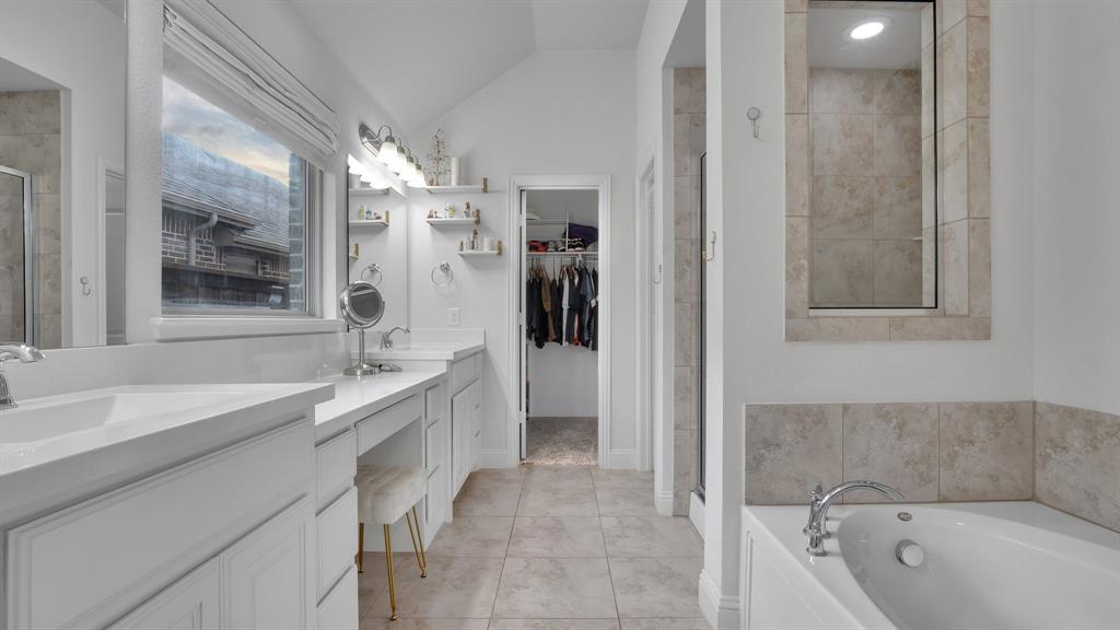 Double vanity with white cabinetry, quartz-surface countertops, soaking tub with tile surround, and neutral-tone tile flooring