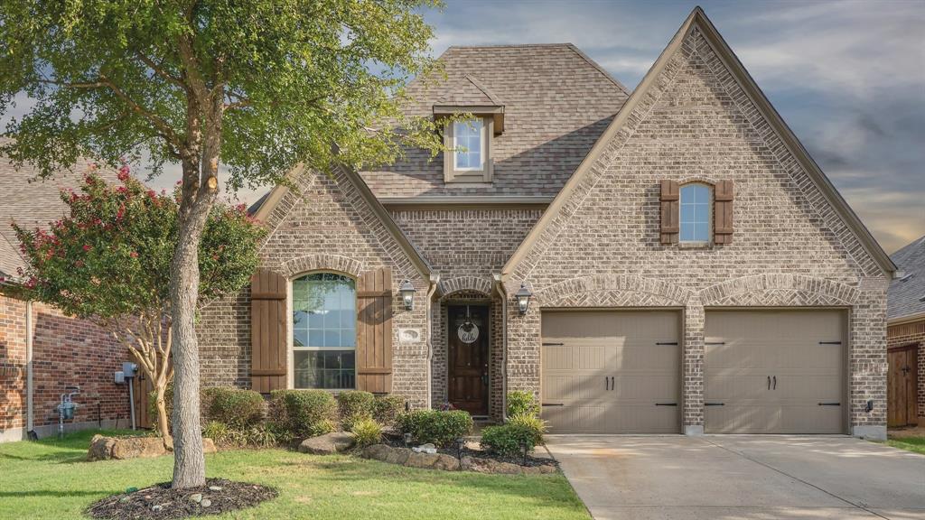 Brick exterior featuring a high-pitched roofline, arched entryway, decorative wood shutters, and an attached two-car garage Brick exterior featuring a high-pitched roofline, arched entryway, decorative wood shutters, and an attached two-car garage