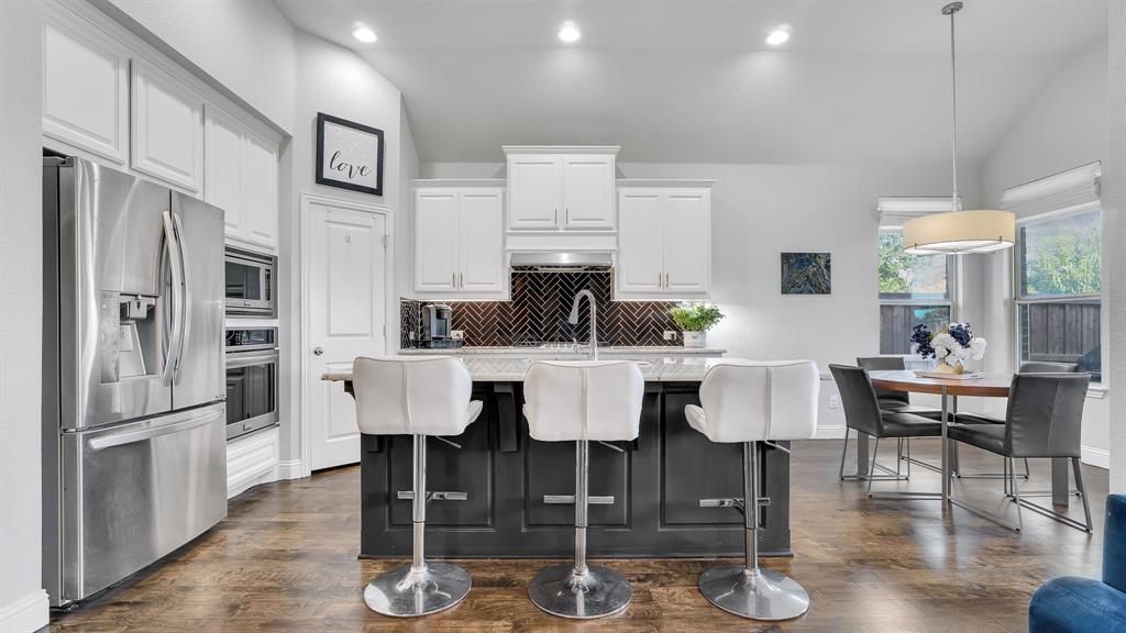 Kitchen featuring a central island with a contrasting dark base, white cabinetry, stainless steel appliances, herringbone-patterned tile backsplash, and wood-finish flooring Kitchen featuring a central island with a contrasting dark base, white cabinetry, stainless steel appliances, herringbone-patterned tile backsplash, and wood-finish flooring