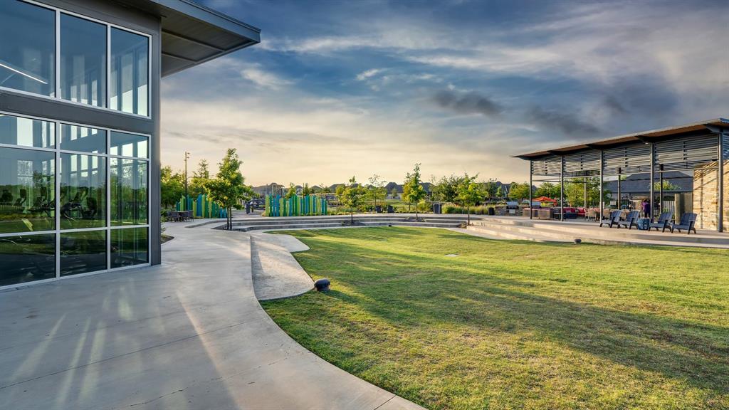 Contemporary multi-purpose common area featuring a floor-to-ceiling glass-walled structure, expansive manicured lawn, tiered concrete amphitheater, and a covered pavilion with metal roof framing