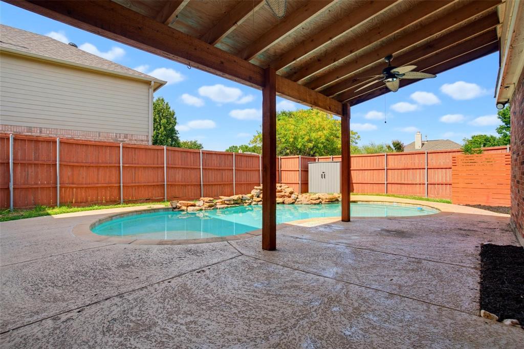 Covered concrete patio with a wooden beam ceiling and ceiling fan, featuring a swimming pool with a rock water feature enclosed by a wooden privacy fence