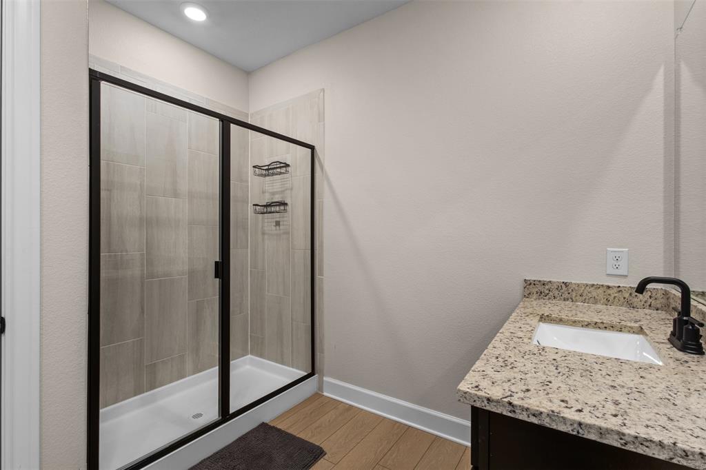 Walk-in shower with floor-to-ceiling tile surround and black metal frame, stone-finish countertop, undermount sink, matte black faucet, and wood-finish flooring
