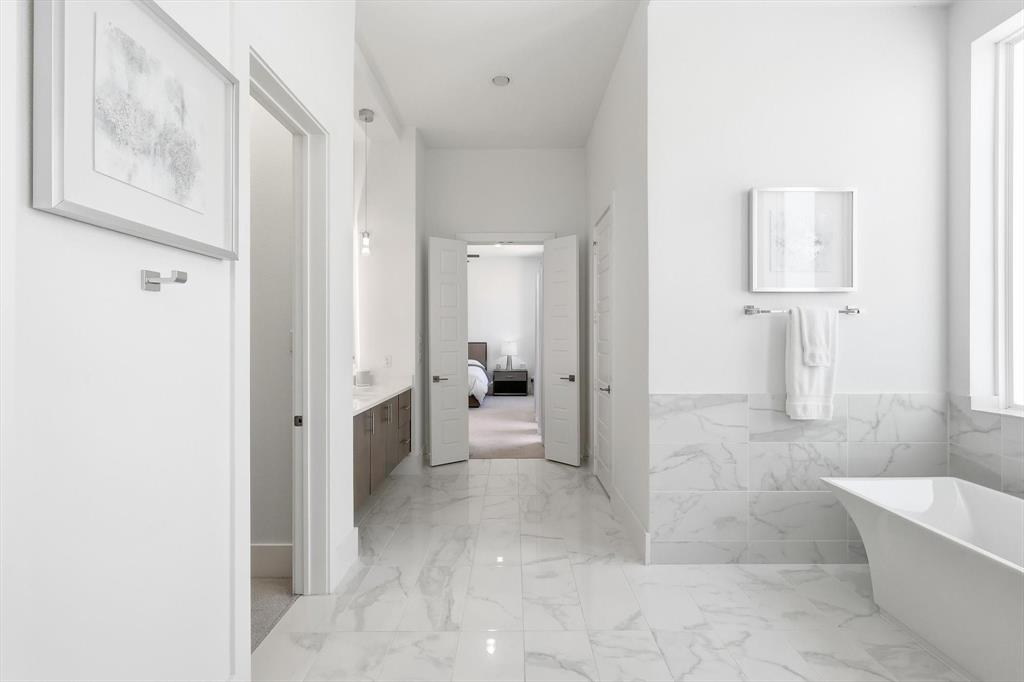 Freestanding soaking tub featuring a stone-look tiled surround, light-toned stone flooring, wall-mounted hardware, and double-door entry