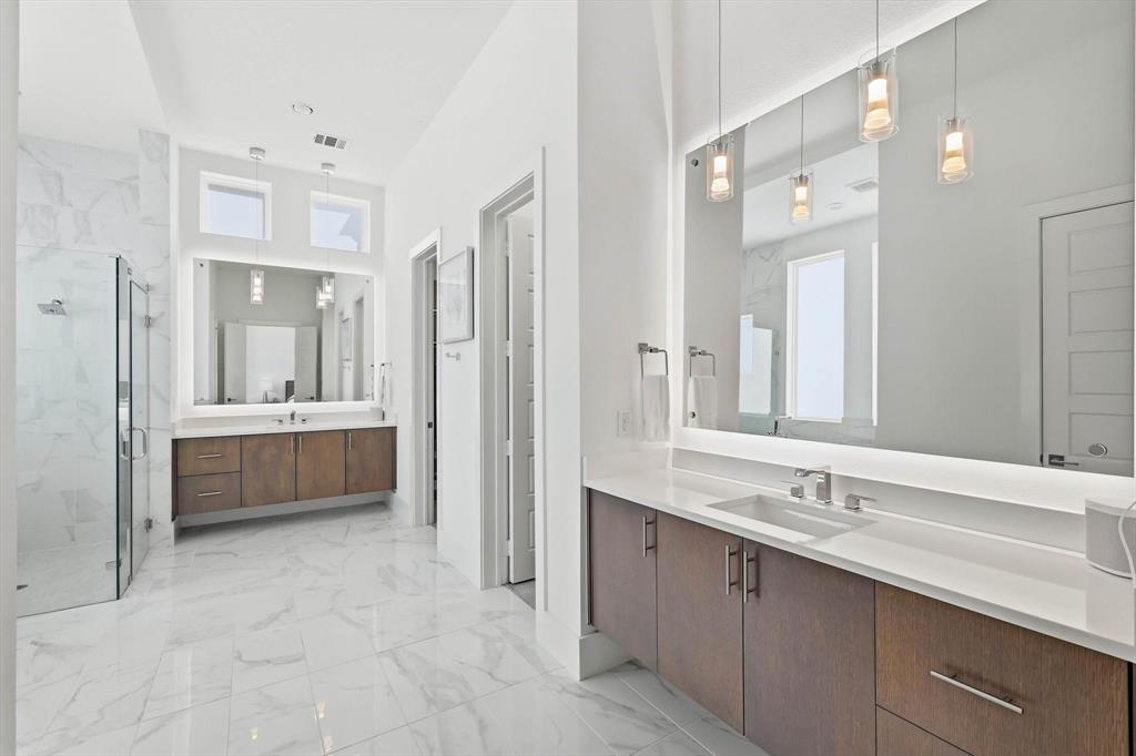 Bathroom featuring dual wood-finish vanities with quartz countertops, large backlit mirrors, pendant lighting, and oversized light-toned tile flooring