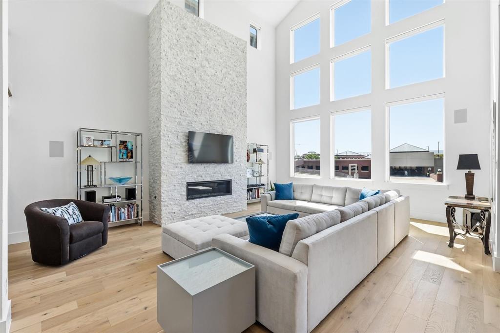 Double-height ceiling, floor-to-ceiling multi-pane windows, and a full-height stone-clad fireplace featuring a linear built-in hearth, complemented by light wood-finish flooring