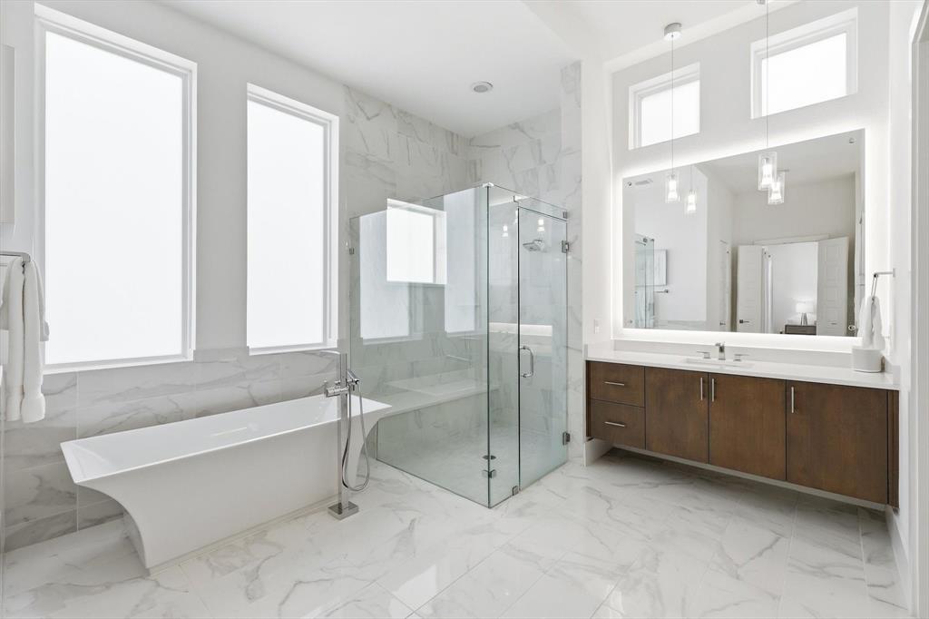 Freestanding soaking tub, glass-enclosed shower with bench seating, dual-sink wood vanity, large backlit mirror, and floor-to-ceiling stone-style wall tile