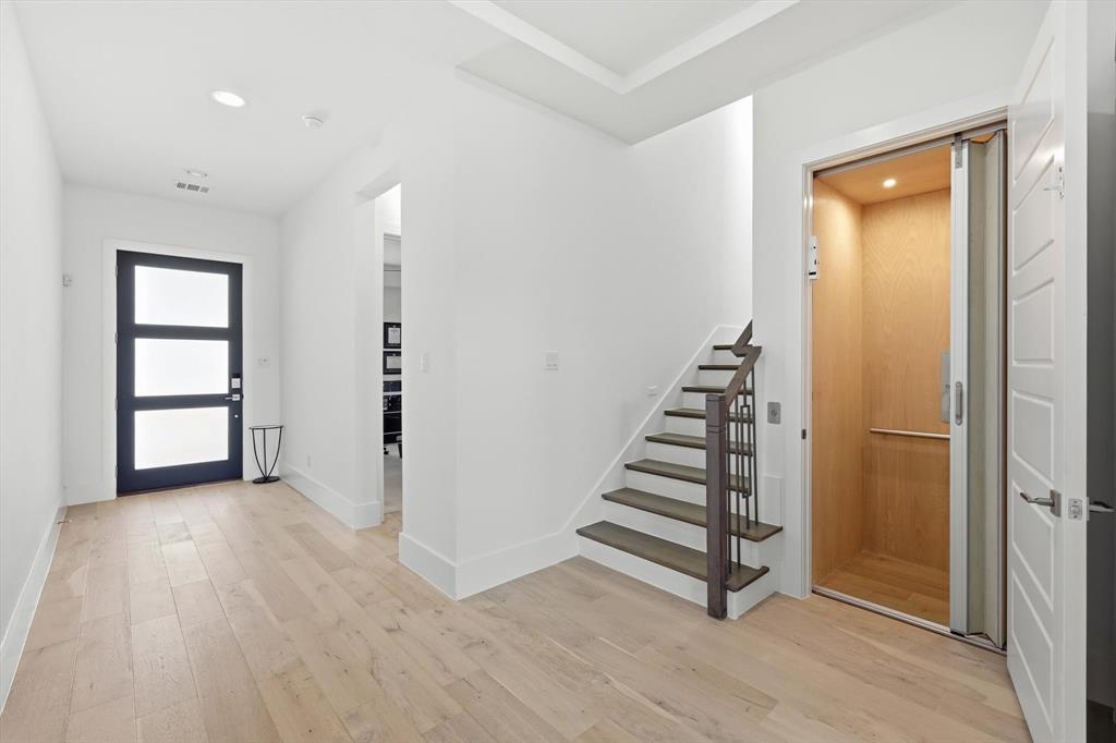 Entryway featuring light-toned wood-finish flooring, a dark-framed multi-pane glass door, recessed ceiling lighting, a staircase with dark wood treads and a coordinating metal railing, and a built-in elevator with wood-panel interior finishes Entryway featuring light-toned wood-finish flooring, a dark-framed multi-pane glass door, recessed ceiling lighting, a staircase with dark wood treads and a coordinating metal railing, and a built-in elevator with wood-panel interior finishes
