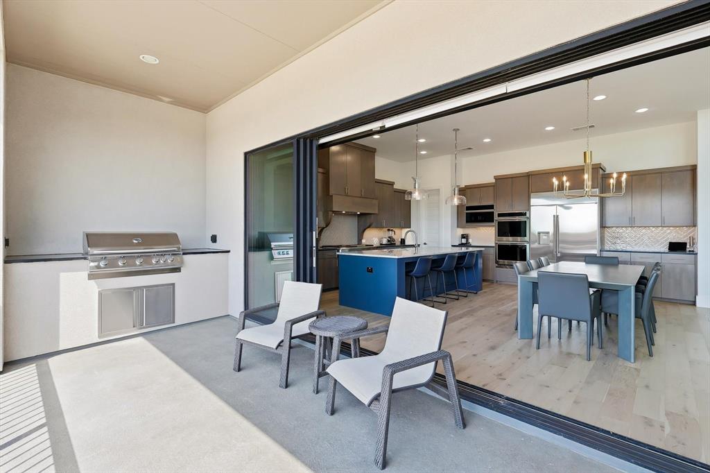 Covered patio featuring a built-in stainless steel grill, retractable glass wall system, wood-finish flooring, central kitchen island with seating, and dual wall-mounted ovens