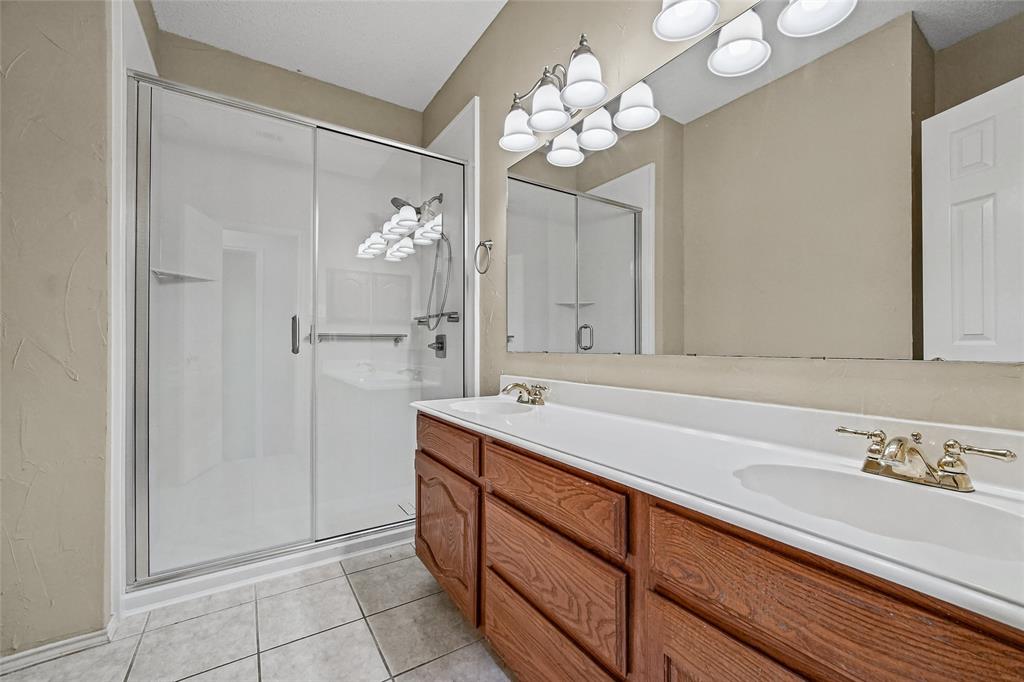 Bathroom featuring a double-sink vanity with wood-finish cabinetry, a full-width wall mirror, a glass-enclosed shower with a sliding door, and neutral tile flooring