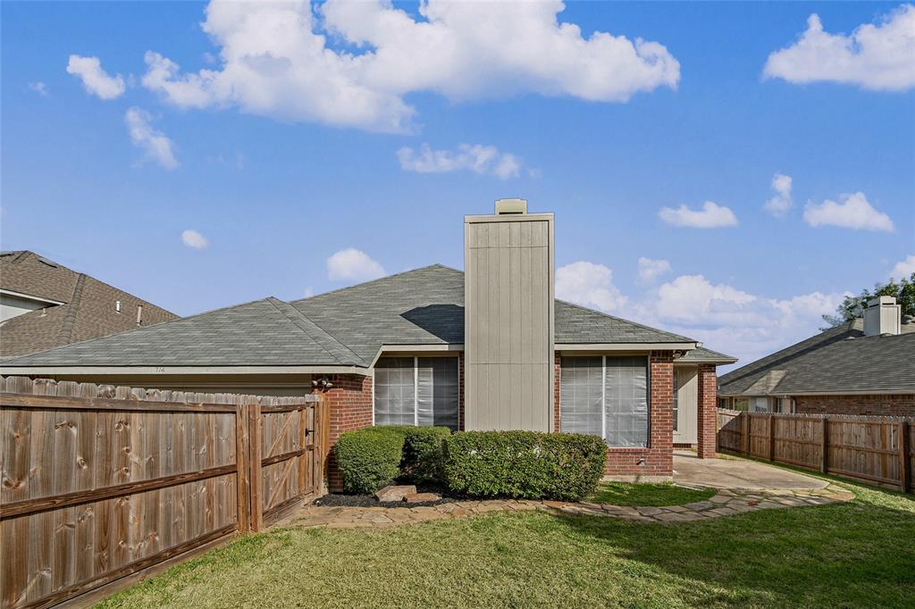 Exterior rear elevation featuring brick siding, a tall chimney, shingle roofing, and a flagstone pathway leading to a concrete patio enclosed by a wood privacy fence