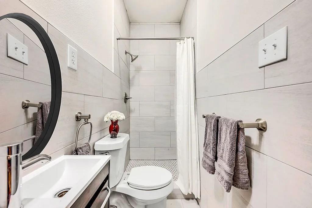 Full-wall horizontal tiling, oval vanity mirror with a black frame, wall-mounted faucet, shower with metallic hardware, and mosaic floor tiling