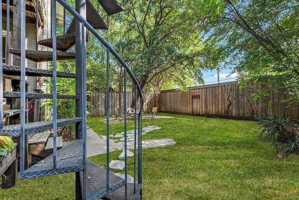 Exterior metal spiral staircase with diamond-plate treads, manicured lawn, stone paver path, and full-height wood privacy fencing