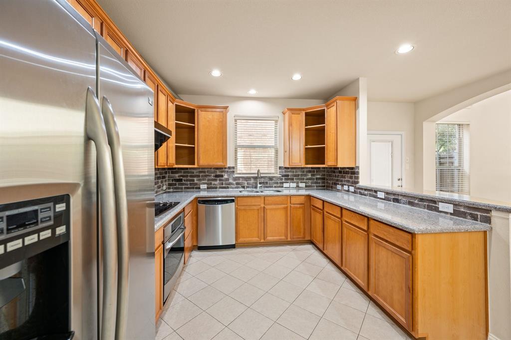 Kitchen featuring wood cabinetry, stone-surface countertops, brick-style tile backsplash, stainless steel appliances, and tile flooring