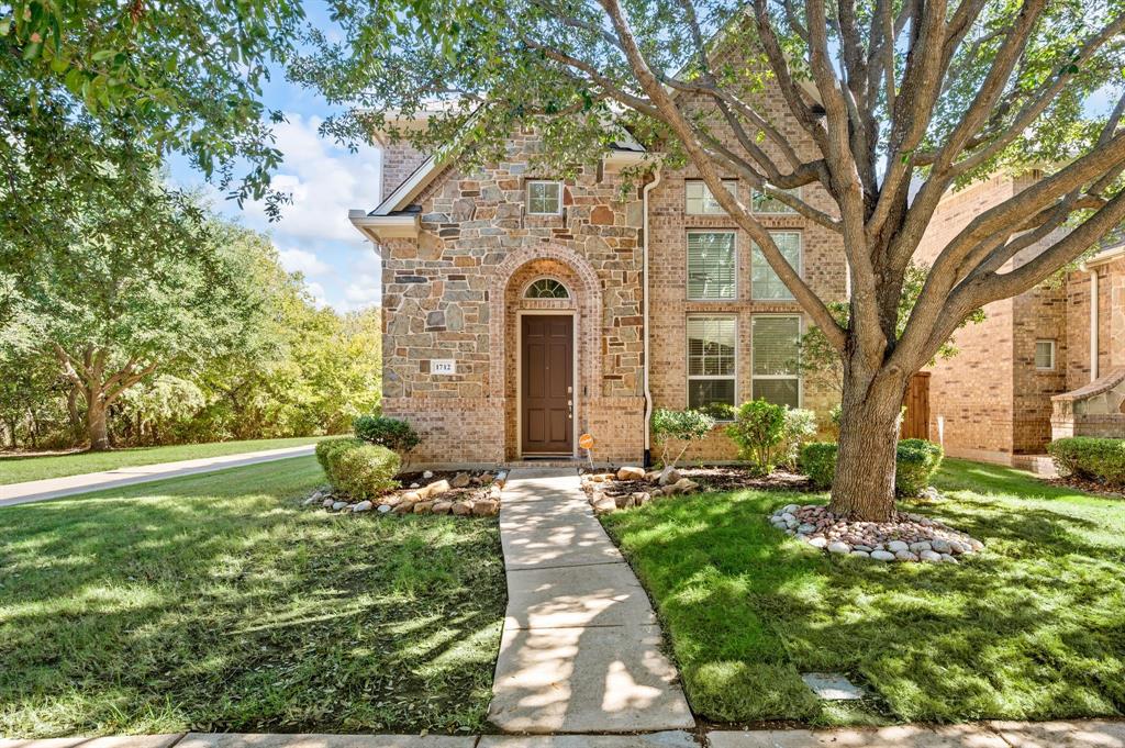 Multi-story stone masonry exterior featuring an arched entryway, tall multi-pane windows, and a mature tree-lined front yard with stone-bordered landscaping