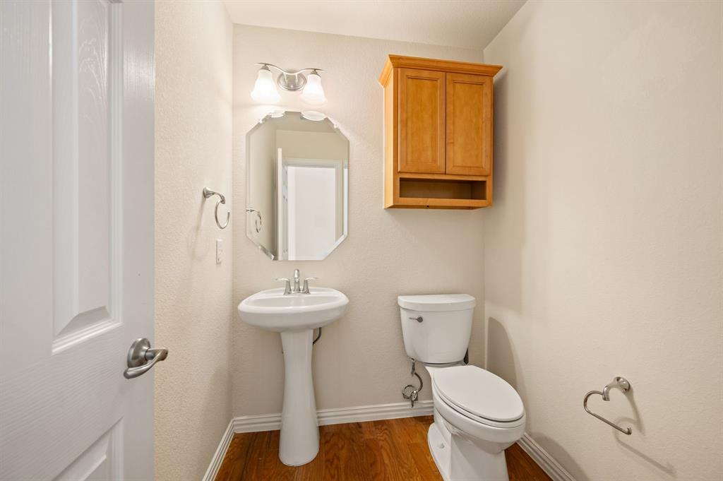 Pedestal sink with polished chrome faucet, octagonal wall mirror, dual-light vanity fixture, wooden wall-mounted storage cabinet, and wood-finish flooring