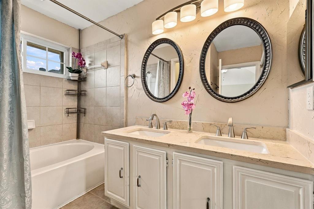 Dual-sink vanity with stone-finish countertop, oval wall-mounted mirrors, light-finish cabinetry with metallic hardware, and a tile-surround soaking tub