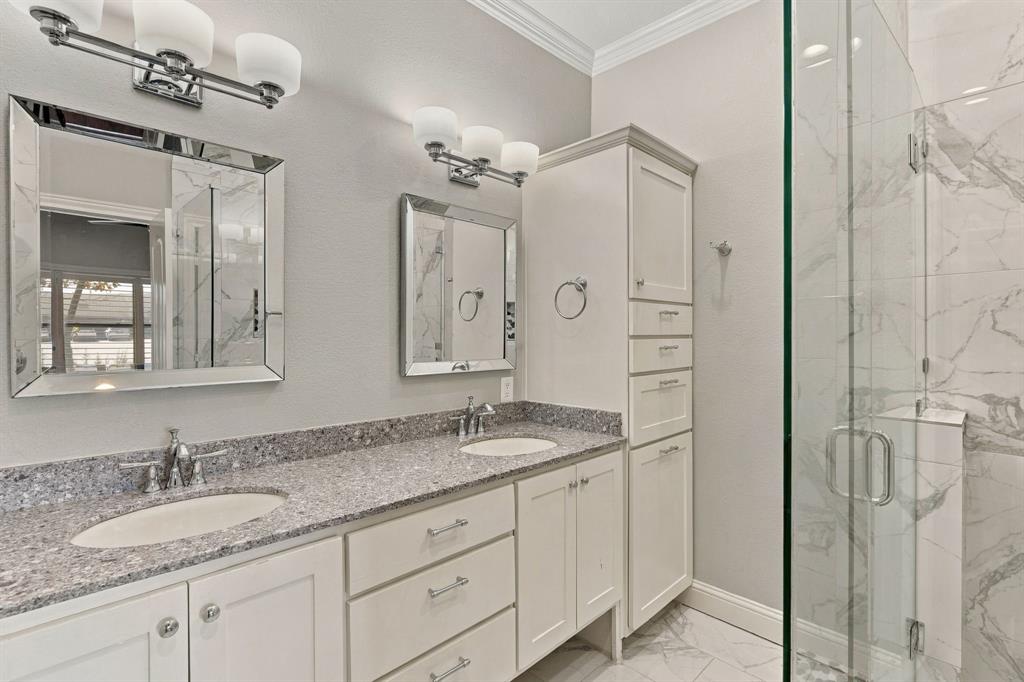 Dual-sink vanity with stone-finish countertop, crisp white cabinetry with floor-to-ceiling storage, framed mirrors, multi-bulb vanity lighting, and walk-in shower with floor-to-ceiling tile surround