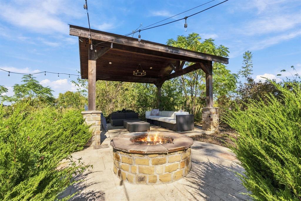 Covered timber-frame pavilion with stone pillar bases, circular stone fire pit, paver patio, and overhead bistro-style lighting