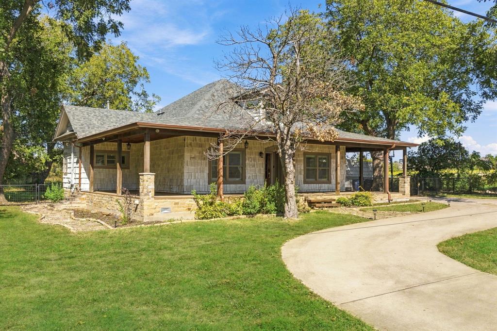 Single-story residence featuring a stone veneer exterior, wraparound covered porch, stone column supports, shingle roof, and a paved driveway Single-story residence featuring a stone veneer exterior, wraparound covered porch, stone column supports, shingle roof, and a paved driveway