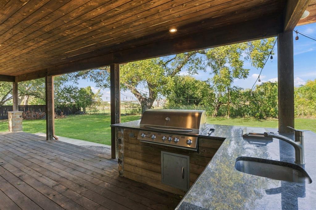 Covered outdoor kitchen featuring wood-plank ceiling, stone-accented base, stainless steel grill, natural stone countertop, and undermount sink