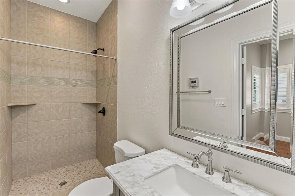 Bathroom featuring a vanity with stone-finish countertop, undermount sink, and integrated mirror, accompanied by a tiled walk-in shower with decorative glass inlay and pebble-texture flooring
