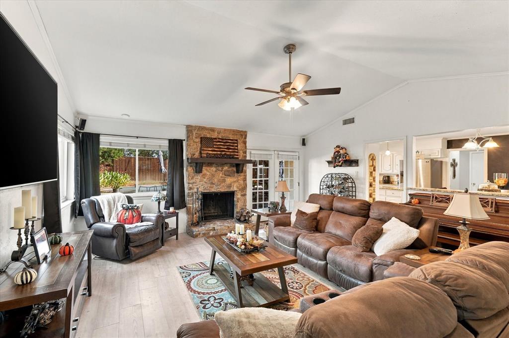 Vaulted ceiling, stone-faced fireplace with a wood mantel, wood-finish flooring, and a ceiling fan with integrated lighting