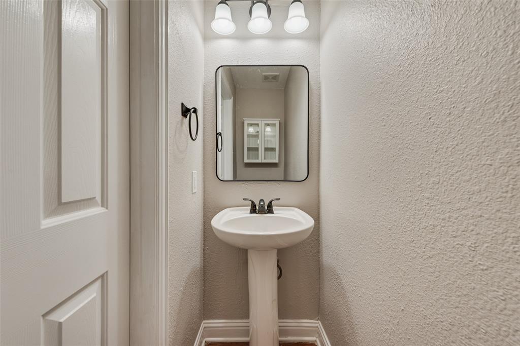 Pedestal sink with a dark metal faucet, rectangular mirror with a black frame, and a three-light fixture with white glass shades