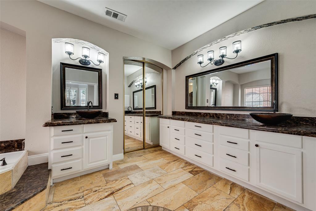 Dual-vanity bathroom featuring white shaker-style cabinetry, dark stone countertops, vessel sinks, framed wall mirrors with integrated lighting, and natural stone flooring
