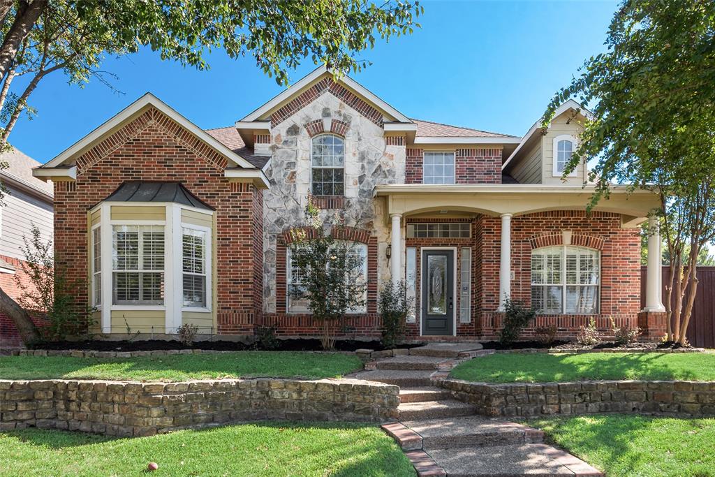 Traditional brick exterior featuring stone veneer accents, a covered front porch with classical columns, a multi-paneled glass front door, and a tiered natural stone retaining wall
