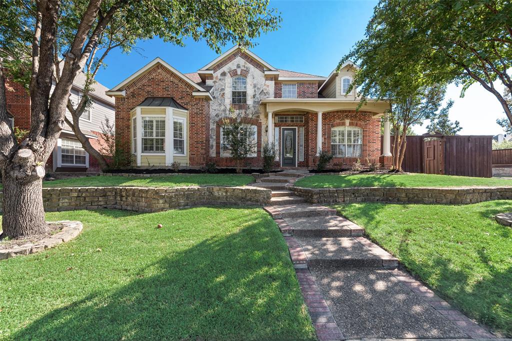 Traditional brick and stone façade featuring a covered front porch with columns, stacked stone retaining walls, and a concrete walkway with brick edging