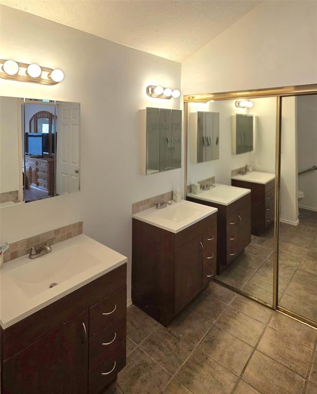 Dual dark-finish vanities with rectangular integrated sinks, stone-finish countertops, tiled backsplashes, and large mirrored closet doors with gold-tone frames over stone-tile flooring