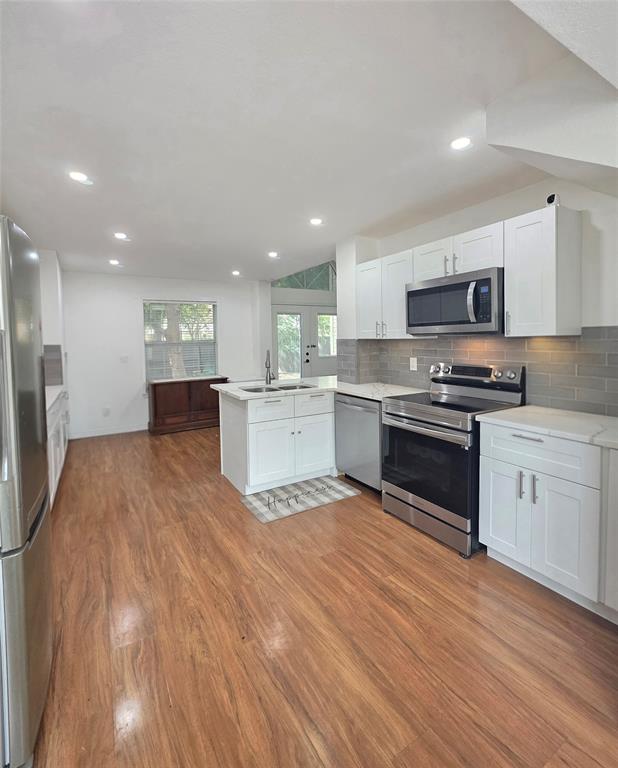 Kitchen featuring wood-finish flooring, white shaker cabinetry, stainless steel appliances, a center island with a dual-basin sink, and gray subway tile backsplash