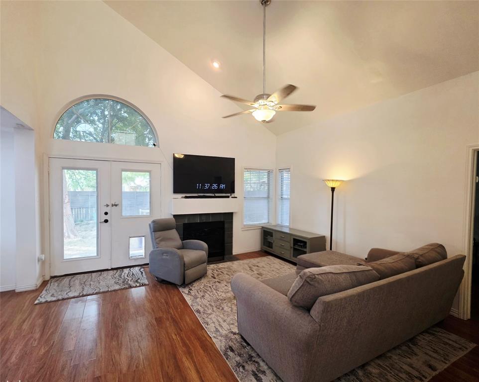 Vaulted ceiling with a central brushed metal ceiling fan, wood-finish flooring, fireplace with a tiled surround and mantel, and a multi-pane arch-top window above a double-entry door