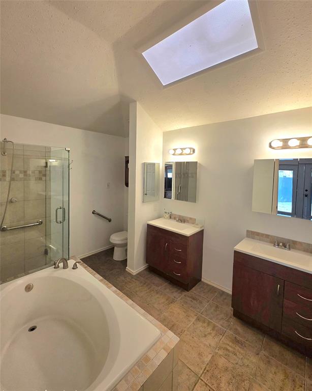 Bathroom featuring a soaking tub, glass-enclosed shower, dual wood-finish vanities with rectangular sinks, and a skylight