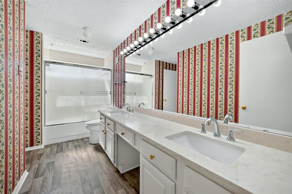 Dual-sink vanity with a light-toned countertop, wood-finish flooring, full-width wall mirror, and tub-shower combination with sliding glass doors