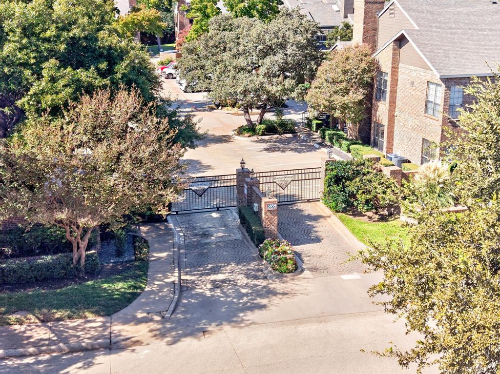 Gated entry featuring a brick-pillared wrought iron gate, paved driveway, mature trees, and landscaping