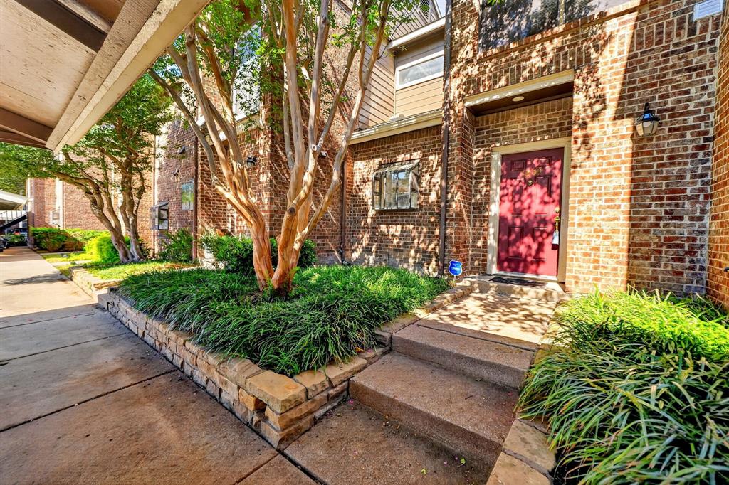 Brick facade exterior featuring a solid red entry door, stacked stone retaining wall, mature crepe myrtle tree, and concrete walkway